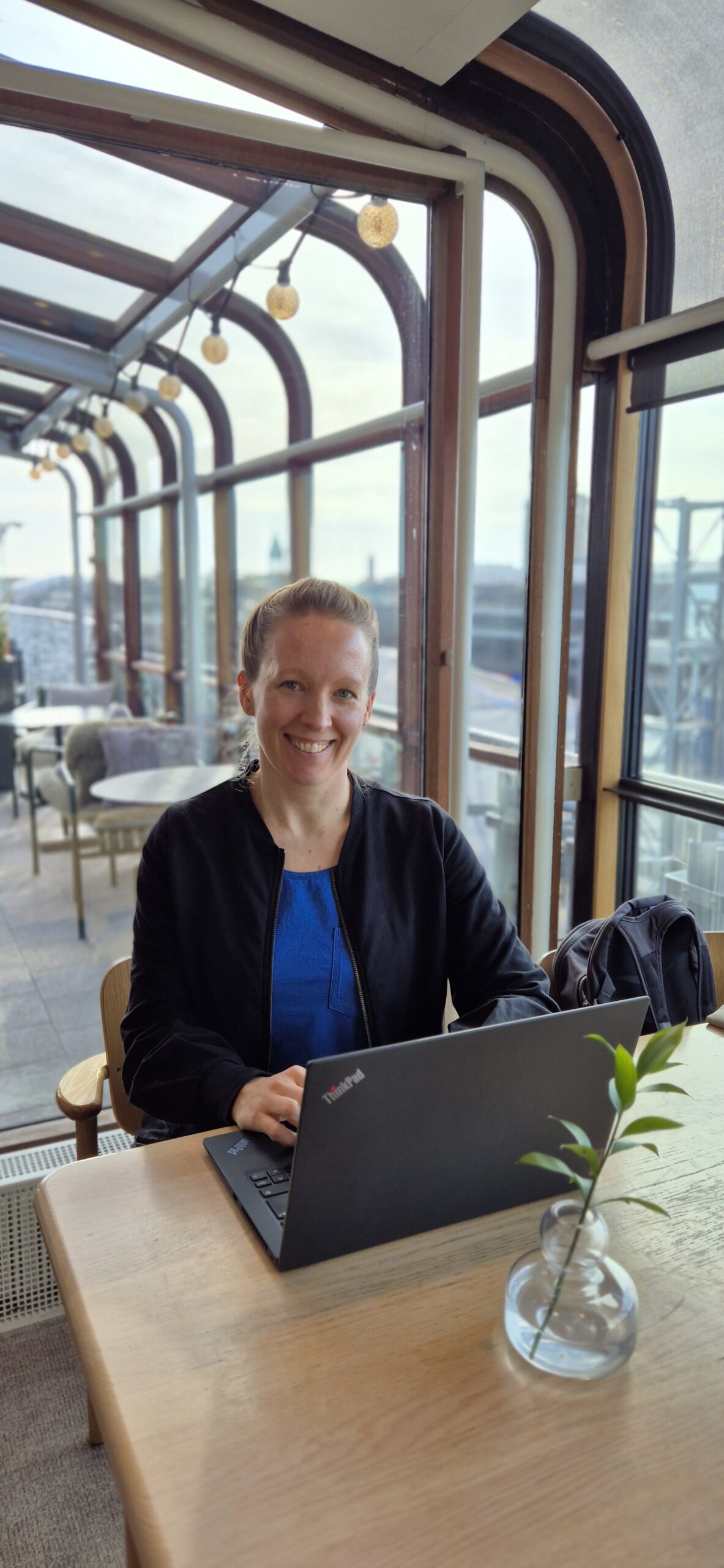 Kuva iloisesta naisesta, yrityksen perustajasta, kannettavan tietokoneen äärellä työskentelemässä. Taustalla suuret ikkunat ja ravintolamiljöö, tietokoneen edessä vihreä oksa maljakossa.