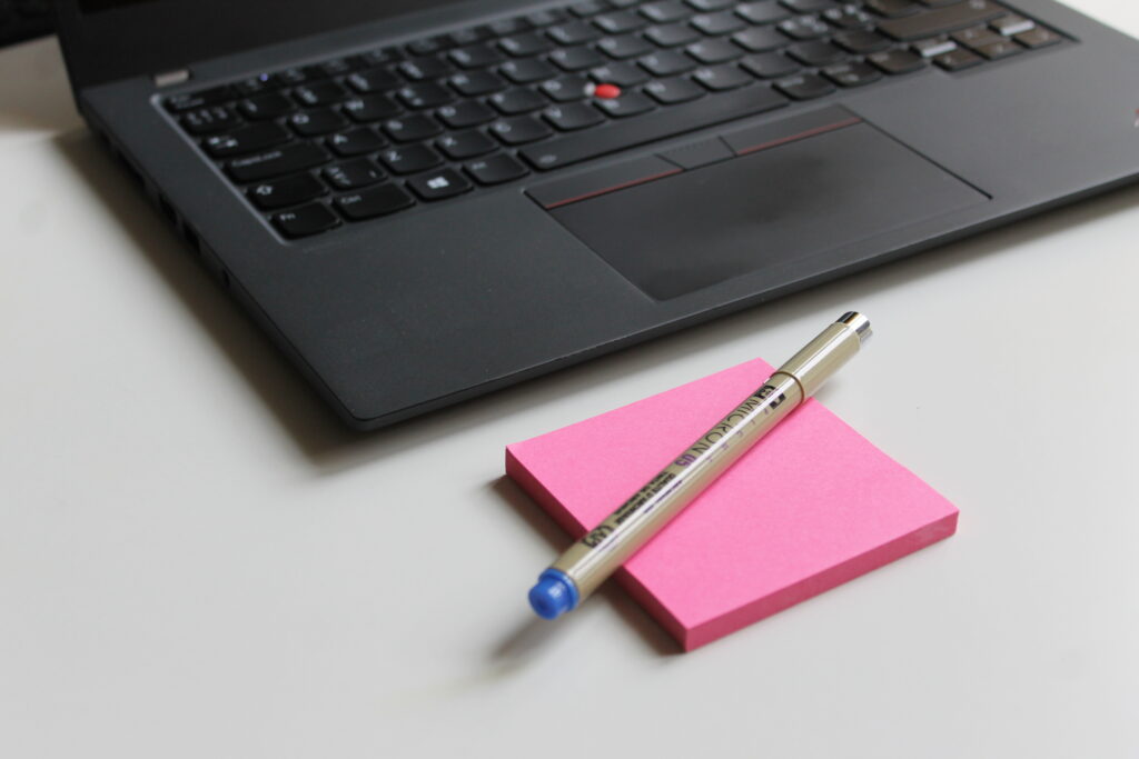 Taustalla kannettavan tietokoneen näppäimistö, etualalla pinkki post-it-nippu jonka päällä kynä. 