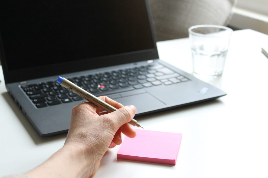 Taustakuva, joka kuvaa toimistotyöskentelyä. Ihmisen käsi ja kädessä kynä, joka kirjoittaa pinkille post-it-lapulle. Takana kannettava tietokone, jonka vieressä vesilasi. 